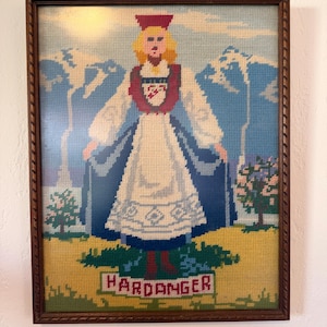 May include: Framed needlepoint artwork depicting a person in traditional Norwegian Hardanger attire. The artwork features a figure in a white dress, red hat, and blue cape, set against a backdrop of mountains and the word "HARDANGER".