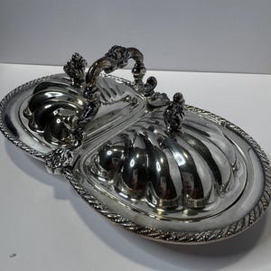 May include: Silver-plated serving dish with two compartments and lids. The oval dish has a decorative rope-like border and a handle. Each lid is topped with a small decorative element. The dish is designed for serving food.