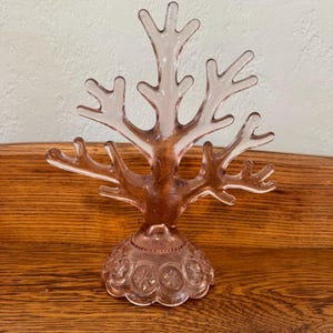 May include: A pale pink glass jewelry stand shaped like a branching tree. The base features a circular design with embossed star-like patterns around the perimeter, providing a stable foundation for the multi-pronged structure above.