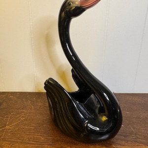 May include: A black ceramic swan figurine with a long, curved neck and a red beak. The swan has a glossy finish and a small opening in the back, possibly for holding flowers or small items. The figurine is a decorative item.