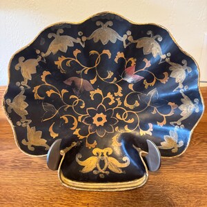 May include: A black decorative dish with a scalloped edge, featuring gold and orange floral designs. The dish is supported by a dark metal stand and sits on a wooden surface. The overall design is ornate and elegant.