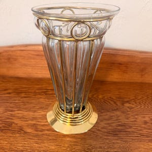May include: A clear glass vase with vertical ribbed detailing and gold-colored metal accents. The vase has a flared shape with a circular base and a decorative rim. The gold accents create a vintage or antique aesthetic.