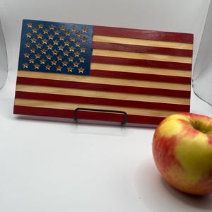 May include: A wooden American flag with a blue field of stars and red and natural wood stripes. The flag is displayed on a black metal stand. A red and yellow apple sits in the foreground.