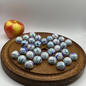 May include: A collection of white marbles with blue and teal swirls, displayed on a round wooden tray. A red and yellow apple sits to the side. The marbles are a classic toy, perfect for games or decorative purposes.
