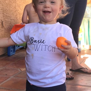 Op de afbeelding: Een jong kind draagt een wit T-shirt met de tekst "Basic Witch" in zwarte letters. Het kind houdt een sinaasappel in zijn rechterhand en glimlacht.