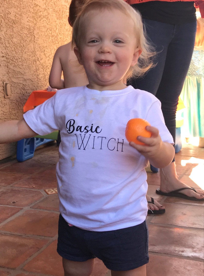 Op de afbeelding: Een jong kind draagt een wit T-shirt met de tekst "Basic Witch" in zwarte letters. Het kind houdt een sinaasappel in zijn rechterhand en glimlacht.