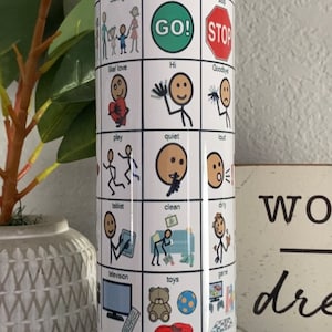 Pode incluir: Uma caneca cilíndrica branca com uma base de plástico transparente. A caneca tem uma grade de imagens e texto, incluindo "bathroom", "yes", "no", "sibling", "go", "stop", "GO!", "STOP", "Hi", "like/love", "Goodbye", "play", "quiet", "loud", "tablet", "clean", "dirty", "television", "toys", e "game".