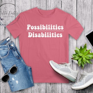 May include: A pink t-shirt with the text "Possibilities Disabilities" in white, with a line separating the words. The shirt is laid out on a wooden surface with a pair of white sneakers, a pair of blue jeans, and a potted succulent.