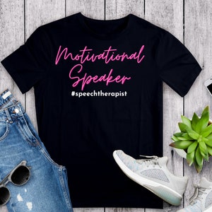 May include: Black t-shirt with pink text that reads "Motivational Speaker #speechtherapist". The shirt is laid flat on a woodgrain surface with a pair of jeans, sunglasses, a plant, and a pair of white sneakers.