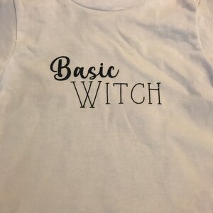 Op de afbeelding: Wit T-shirt met zwarte tekst die "Basic Witch" zegt.