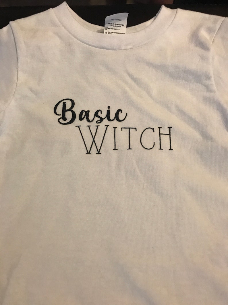 Op de afbeelding: Wit T-shirt met zwarte tekst die "Basic Witch" zegt.