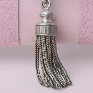 May include: A silver-toned metal tassel pendant featuring a rounded cap with vertical ridges and a series of hanging, flexible chain strands. The pendant has a polished metallic finish and a small circular loop at the top for attachment.