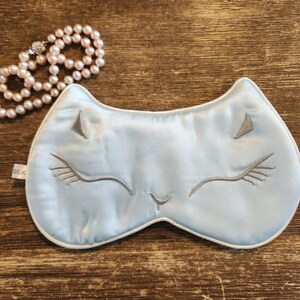 May include: A light blue sleep mask with a white trim and a grey embroidered cat face. The cat has closed eyes and a small nose.