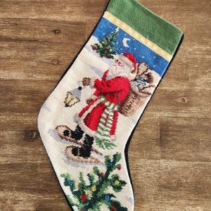 May include: Needlepoint Christmas stocking depicting Santa Claus with a sack of toys. The stocking has a green cuff, black trim, and a winter scene with a Christmas tree and a crescent moon. Festive decor.
