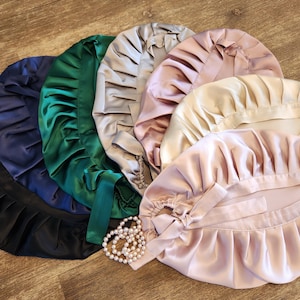 Puede incluir: Un conjunto de seis gorros para dormir de satén en varios colores, incluyendo negro, azul marino, verde, plateado, rosa y beige. Los gorros tienen un diseño fruncido y con volantes y se atan con una cinta.