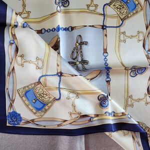 May include: A folded silk scarf with a repeating equestrian-themed pattern. The design features images of horse bits, bridles, and saddlebags in shades of brown, blue, and gold against a cream and light blue background. The scarf has a dark blue border.