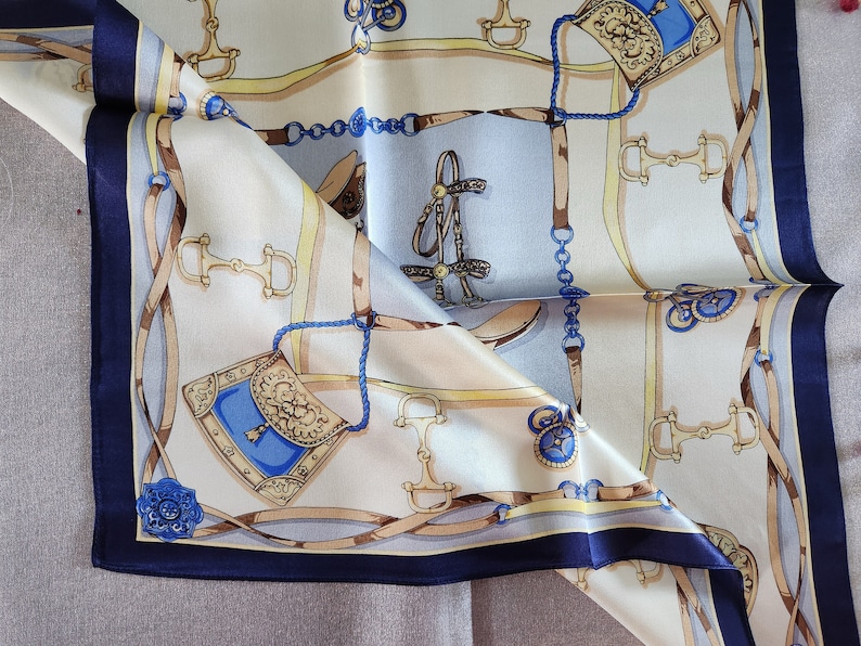 May include: A folded silk scarf with a repeating equestrian-themed pattern. The design features images of horse bits, bridles, and saddlebags in shades of brown, blue, and gold against a cream and light blue background. The scarf has a dark blue border.