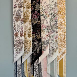 May include: A collection of seven silk scarves with floral patterns. Each scarf features a different color scheme, including white, black, and gold, with detailed botanical designs. The scarves are hanging on a white hanger against a light blue wall.