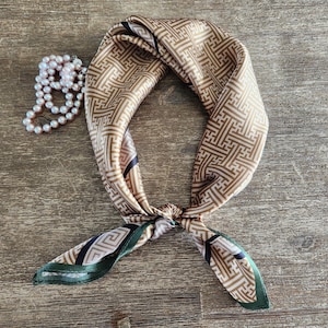 May include: A tan and brown patterned silk scarf tied in a knot, with green and black accents on the ends. A strand of white pearls is next to the scarf. The scarf has a geometric design.