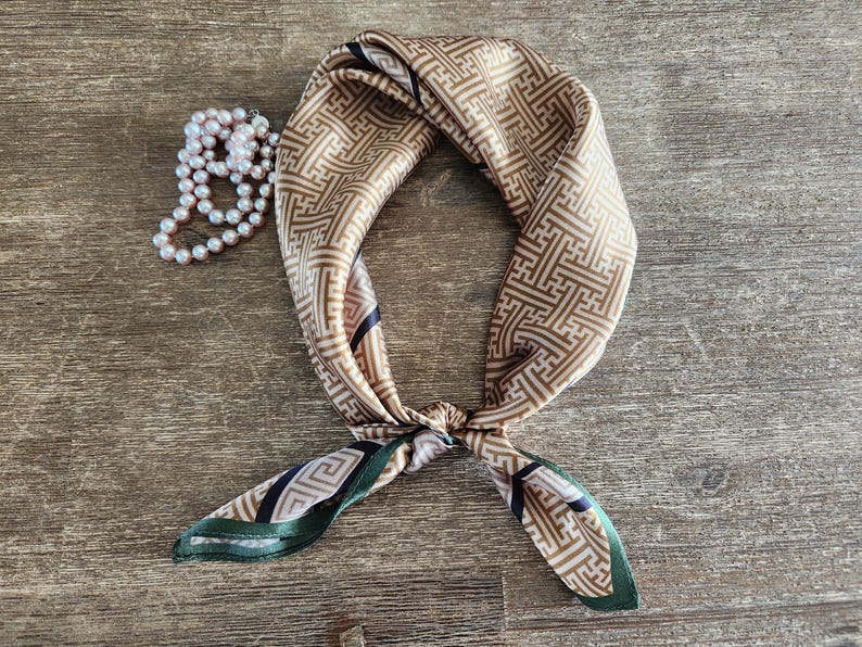 May include: A tan and brown patterned silk scarf tied in a knot, with green and black accents on the ends. A strand of white pearls is next to the scarf. The scarf has a geometric design.