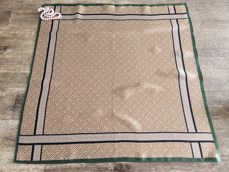 May include: A tan and cream-colored silk scarf with a geometric pattern. The scarf has a dark green border and black and cream-colored trim. A string of pearls is in the upper left corner. The scarf is approximately 28 inches square.