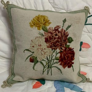 May include: Square decorative cushion with a floral needlepoint design. The cushion features a bouquet of yellow, red, and white flowers with green leaves. It has a light green border with gold trim and decorative tassels at each corner.