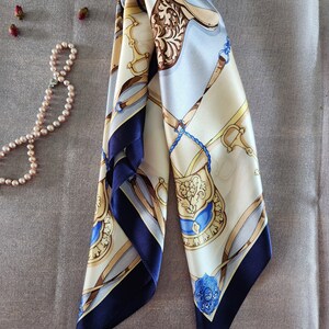 May include: A folded silk scarf with a horse-themed design in cream, light blue, and brown. The scarf features navy blue borders and intricate patterns, with gold and blue accents. A pearl necklace and dried rosebuds are in the background.