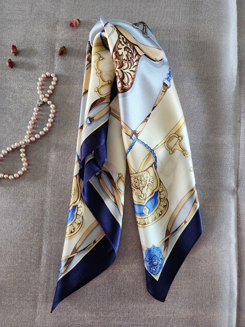 May include: A folded silk scarf with a horse-themed design in cream, light blue, and brown. The scarf features navy blue borders and intricate patterns, with gold and blue accents. A pearl necklace and dried rosebuds are in the background.