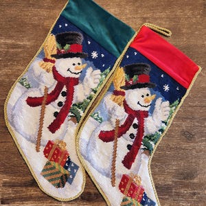 K&ouml;nnte beinhalten: Zwei Weihnachtsstr&uuml;mpfe mit einem Schneemann-Motiv. Die Str&uuml;mpfe sind aus wei&szlig;em Stoff mit einem gr&uuml;nen und roten Samt-Oberteil. Der Schneemann h&auml;lt einen Besen und tr&auml;gt einen roten Schal und einen schwarzen Zylinder. Die Str&uuml;mpfe sind mit goldener Litze verziert.