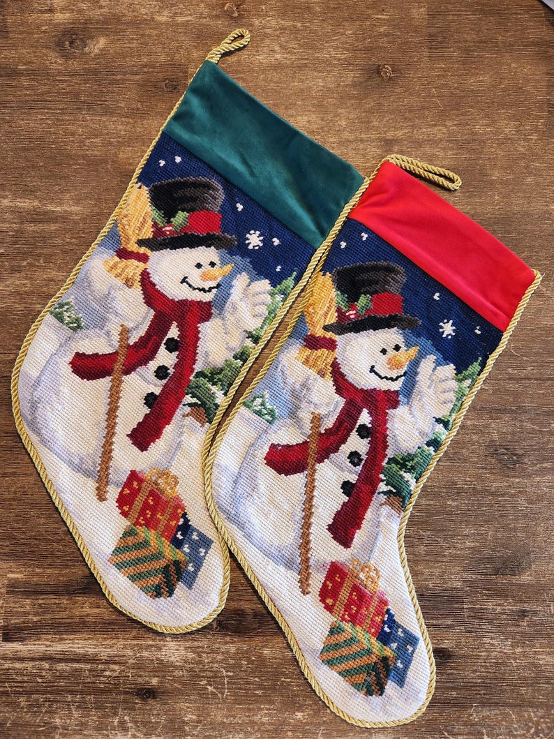 K&ouml;nnte beinhalten: Zwei Weihnachtsstr&uuml;mpfe mit einem Schneemann-Motiv. Die Str&uuml;mpfe sind aus wei&szlig;em Stoff mit einem gr&uuml;nen und roten Samt-Oberteil. Der Schneemann h&auml;lt einen Besen und tr&auml;gt einen roten Schal und einen schwarzen Zylinder. Die Str&uuml;mpfe sind mit goldener Litze verziert.