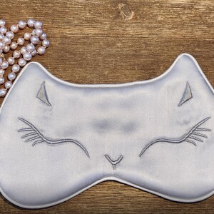 May include: A grey silk sleep mask with a cat face embroidered on it. The mask has a small tag that says "SILK".
