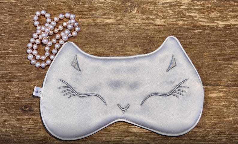 May include: A grey silk sleep mask with a cat face embroidered on it. The mask has a small tag that says "SILK".