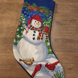 Puede incluir: Calcetín navideño con un diseño detallado de punto de aguja. El calcetín presenta un muñeco de nieve con gorro y bufanda rojos, rodeado de cardinales y un paisaje nevado. La parte superior es azul con piñas.
