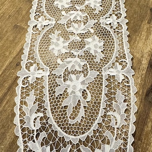 May include: An elongated white lace table runner featuring a repeating floral pattern with intricate petal details and a hexagonal mesh background. The edges of the runner are scalloped, following the curves of the floral motifs throughout the design.
