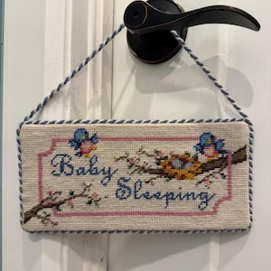 May include: A rectangular needlepoint sign with a cream background, featuring the text "Baby Sleeping" in blue script. The design includes two blue birds, a brown tree branch, and a bird's nest with eggs, all framed by a pink border and blue twisted cord.