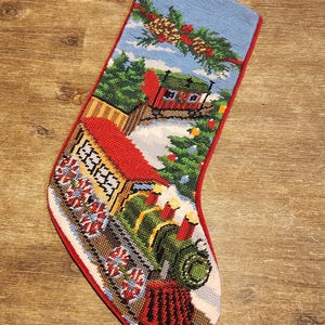 May include: Needlepoint Christmas stocking with a festive train scene. The stocking features a blue sky, green trees, and a red train with white and red accents. The stocking is trimmed in red.