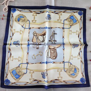 May include: Square silk scarf with a navy blue border. The scarf features a repeating equestrian design in cream, tan, and blue, including saddles, bridles, and decorative elements. The scarf is folded and displayed on a light-colored surface.