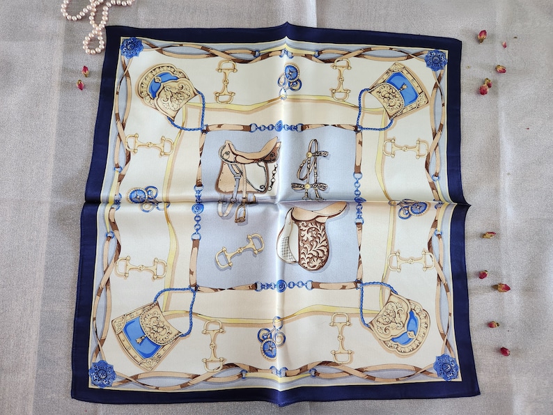 May include: Square silk scarf with a navy blue border. The scarf features a repeating equestrian design in cream, tan, and blue, including saddles, bridles, and decorative elements. The scarf is folded and displayed on a light-colored surface.