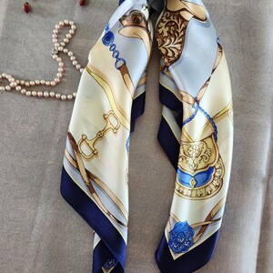 May include: A square silk scarf with a horse-themed design. The scarf features a cream-colored background with blue, gold, and brown equestrian motifs. The edges are a deep navy blue, and a pearl necklace and dried rosebuds are in the background.