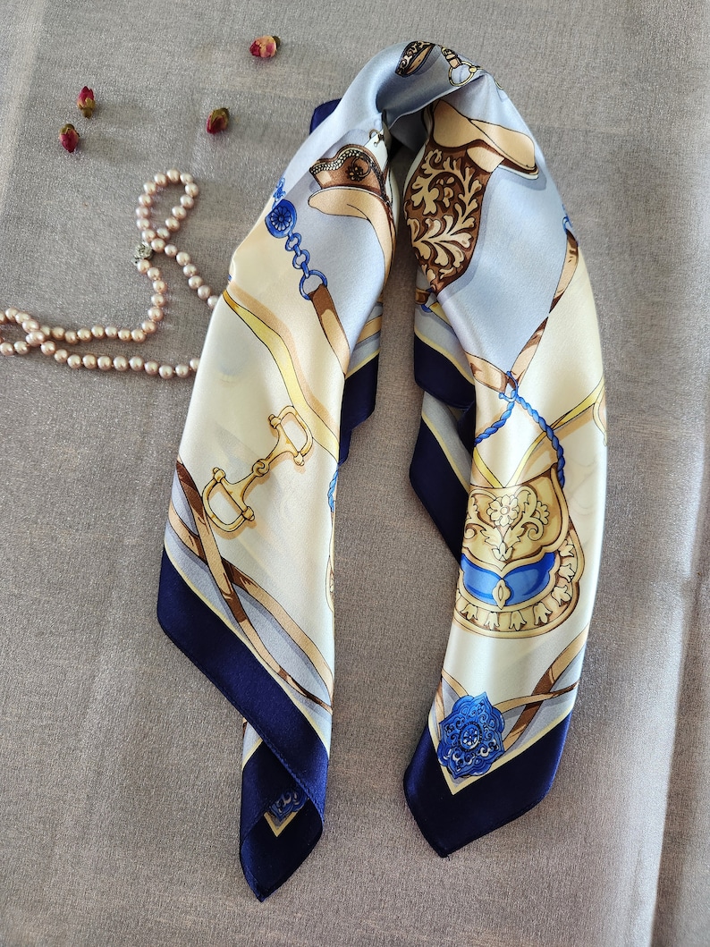 May include: A square silk scarf with a horse-themed design. The scarf features a cream-colored background with blue, gold, and brown equestrian motifs. The edges are a deep navy blue, and a pearl necklace and dried rosebuds are in the background.