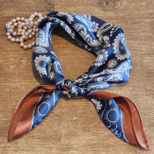 May include: A blue paisley patterned silk scarf with a brown border. The scarf is tied in a knot and the ends are hanging down.