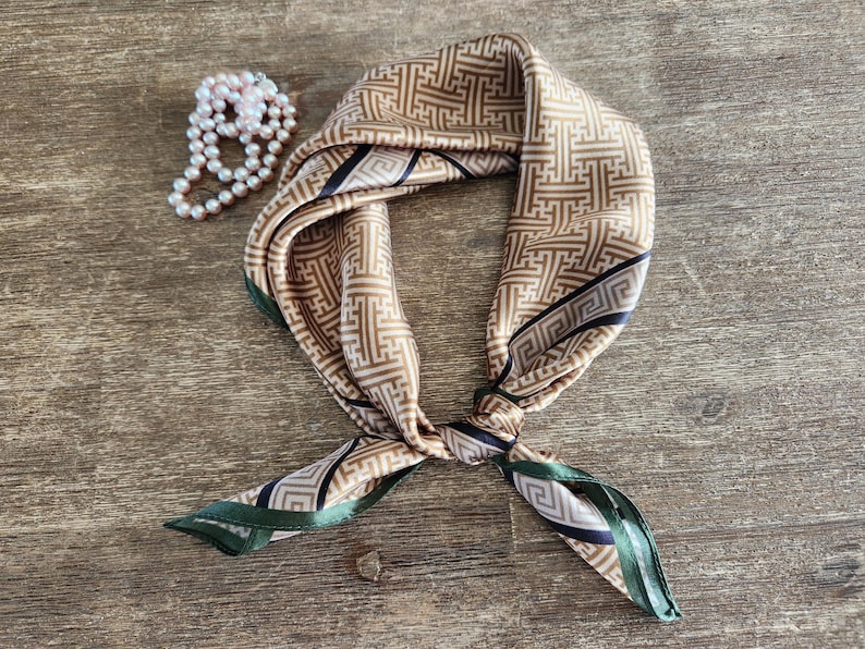 May include: A silk scarf with a geometric pattern in shades of tan and brown, accented with dark green and navy blue borders. A pearl necklace is next to the scarf. The scarf is knotted and displayed on a wooden surface.