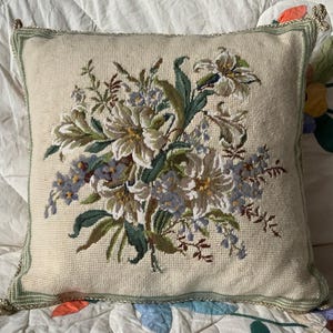 May include: Square cushion with a floral tapestry design. The cushion has a cream background with an embroidered bouquet of white, blue, and green flowers. It is trimmed with a green border and tassels.
