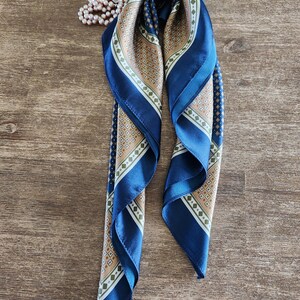May include: A folded blue and tan silk scarf with a geometric pattern and a pearl necklace on a wooden surface. The scarf has a dark blue border and a repeating diamond design in tan, white, and green.