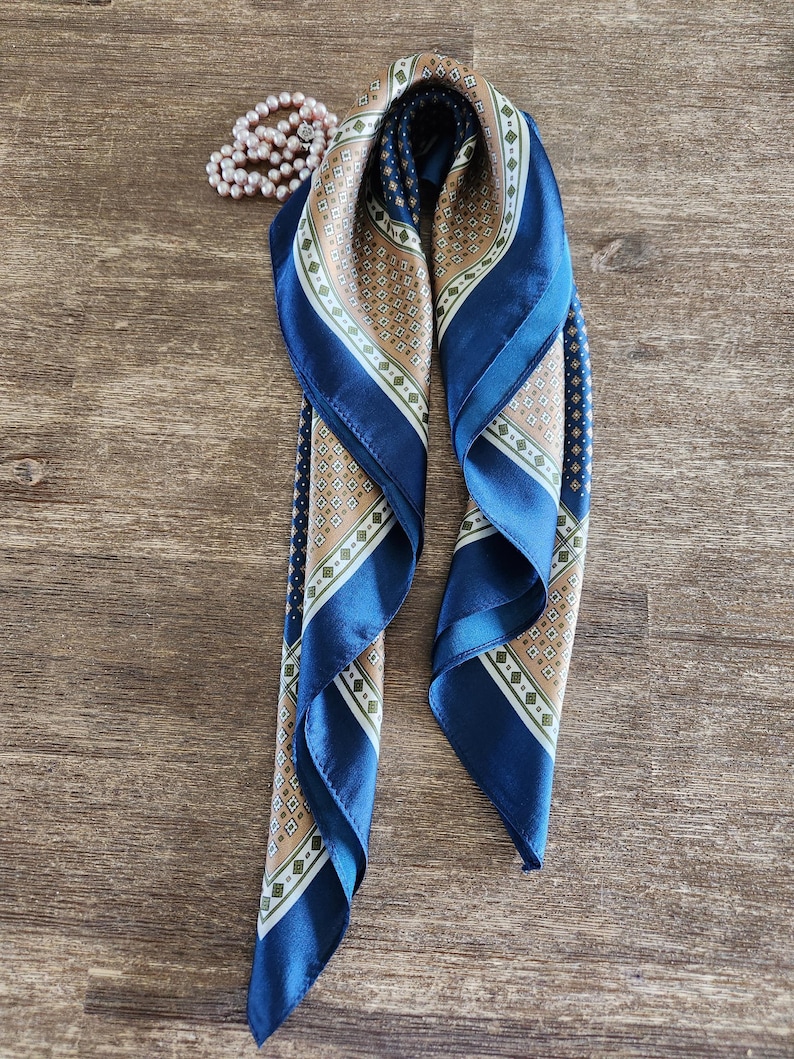 May include: A folded blue and tan silk scarf with a geometric pattern and a pearl necklace on a wooden surface. The scarf has a dark blue border and a repeating diamond design in tan, white, and green.