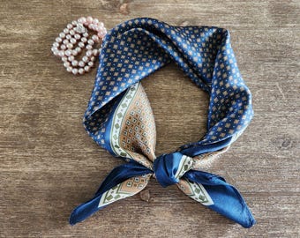 May include: A navy blue silk scarf with a geometric pattern in gold and white. The scarf is knotted and lies on a wooden surface, next to a string of pink pearls. The scarf has a square shape.
