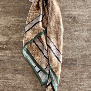 May include: A tan and black patterned silk scarf with a green border. The scarf is draped over a wooden surface. A string of pearls is visible in the upper left corner. The scarf's design features geometric shapes.