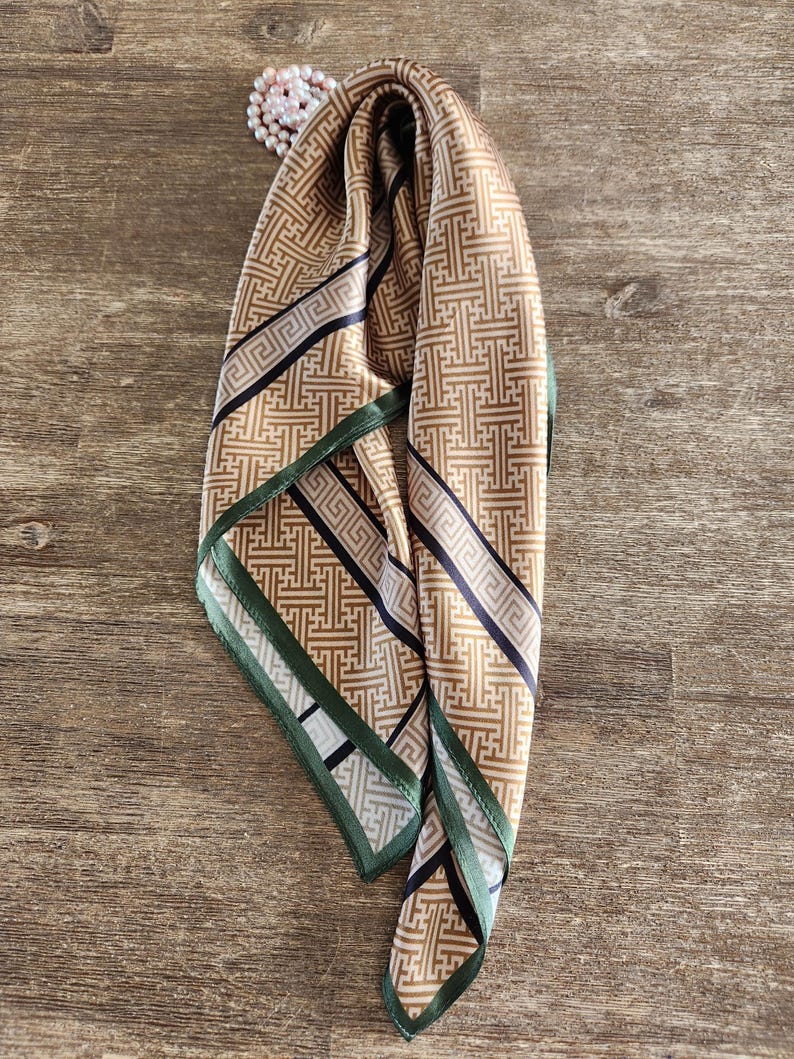 May include: A tan and black patterned silk scarf with a green border. The scarf is draped over a wooden surface. A string of pearls is visible in the upper left corner. The scarf's design features geometric shapes.