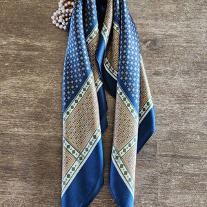 May include: A folded navy blue silk scarf with a geometric pattern in tan, white, and green. The scarf has a dark blue border and is draped on a wooden surface. A string of pearls is visible in the background.
