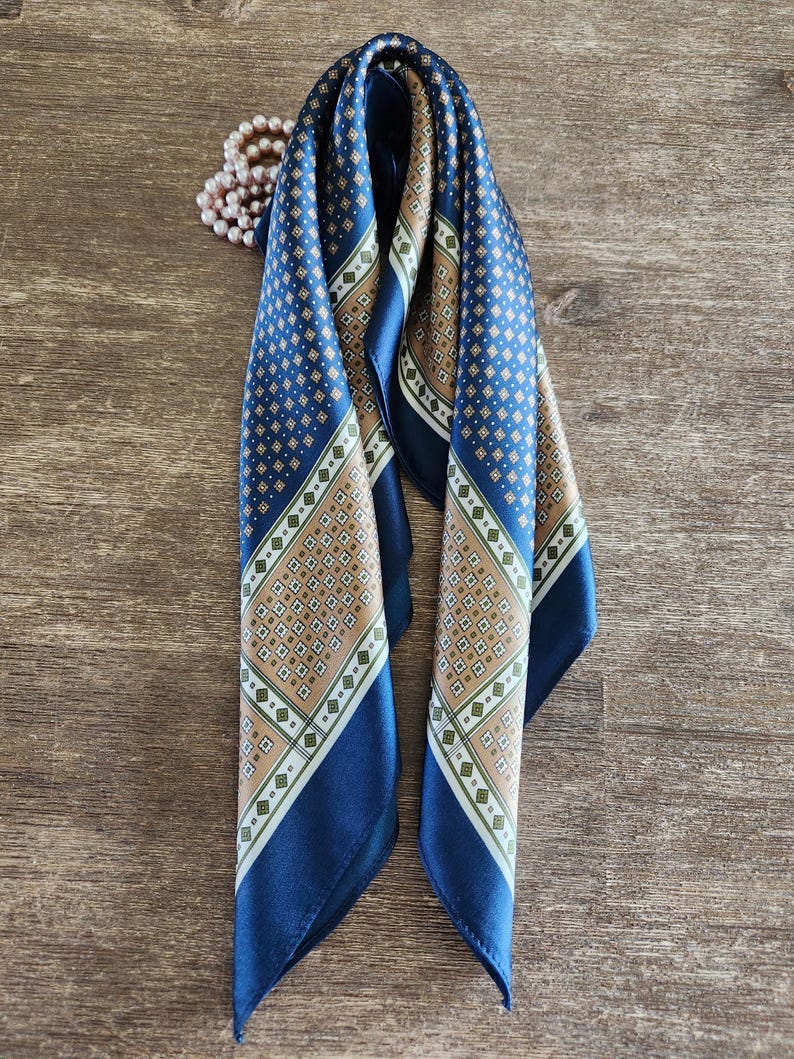 May include: A folded navy blue silk scarf with a geometric pattern in tan, white, and green. The scarf has a dark blue border and is draped on a wooden surface. A string of pearls is visible in the background.